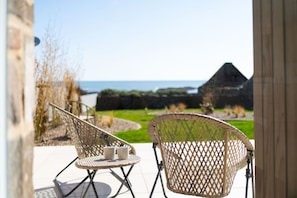 Un patio con sillas de mimbre y una mesa, con vistas a un área verde y una estructura de piedra junto al mar.