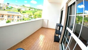 A balcony with a tiled floor, a black metal shelf, and a window with a view of a hillside. - Lisbon, modern T2 with balcony in Barcarena (Oeiras)