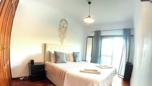 A bedroom with a bed, bedside tables, a nightstand, a dreamcatcher, a hanging light, and a window with curtains. - Lisbon, modern T2 with balcony in Barcarena (Oeiras)