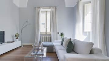 Ein modernes Wohnzimmer mit einer Couch, einem Couchtisch und einem Fernseher auf einem weißen Schrank. Es gibt ein Fenster mit weißen Vorhängen, eine Heizung und eine Pflanze auf dem Boden.