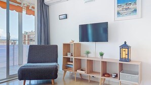 Een moderne woonkamer met een grijze fauteuil, een flatscreen televisie, een houten tv-meubel met decoratieve accessoires, en een stadszicht door grote ramen.