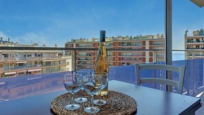 Een tafel met een fles en glazen op een geweven onderzetter, met uitzicht op een stadsgezicht met gebouwen en balkons.