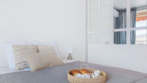 Een slaapkamer met een bed, een nachtkastje met een lamp, een raam met gordijnen en een rieten mandje op het bed.