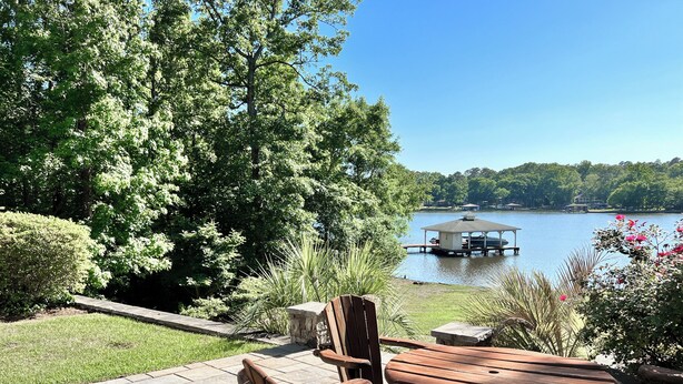 A patio with wooden furniture overlooking a lake, surrounded by lush greenery. - Lakefront Escape on Lake Sinclair • Hot Tub • Ideal for Families & Friends (Eatonton)