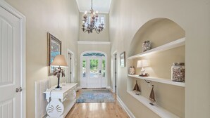 A hallway with wooden flooring, white walls, a chandelier, a door with glass panels, and shelves with decorative items. - Lakefront Escape on Lake Sinclair • Hot Tub • Ideal for Families & Friends (Eatonton)
