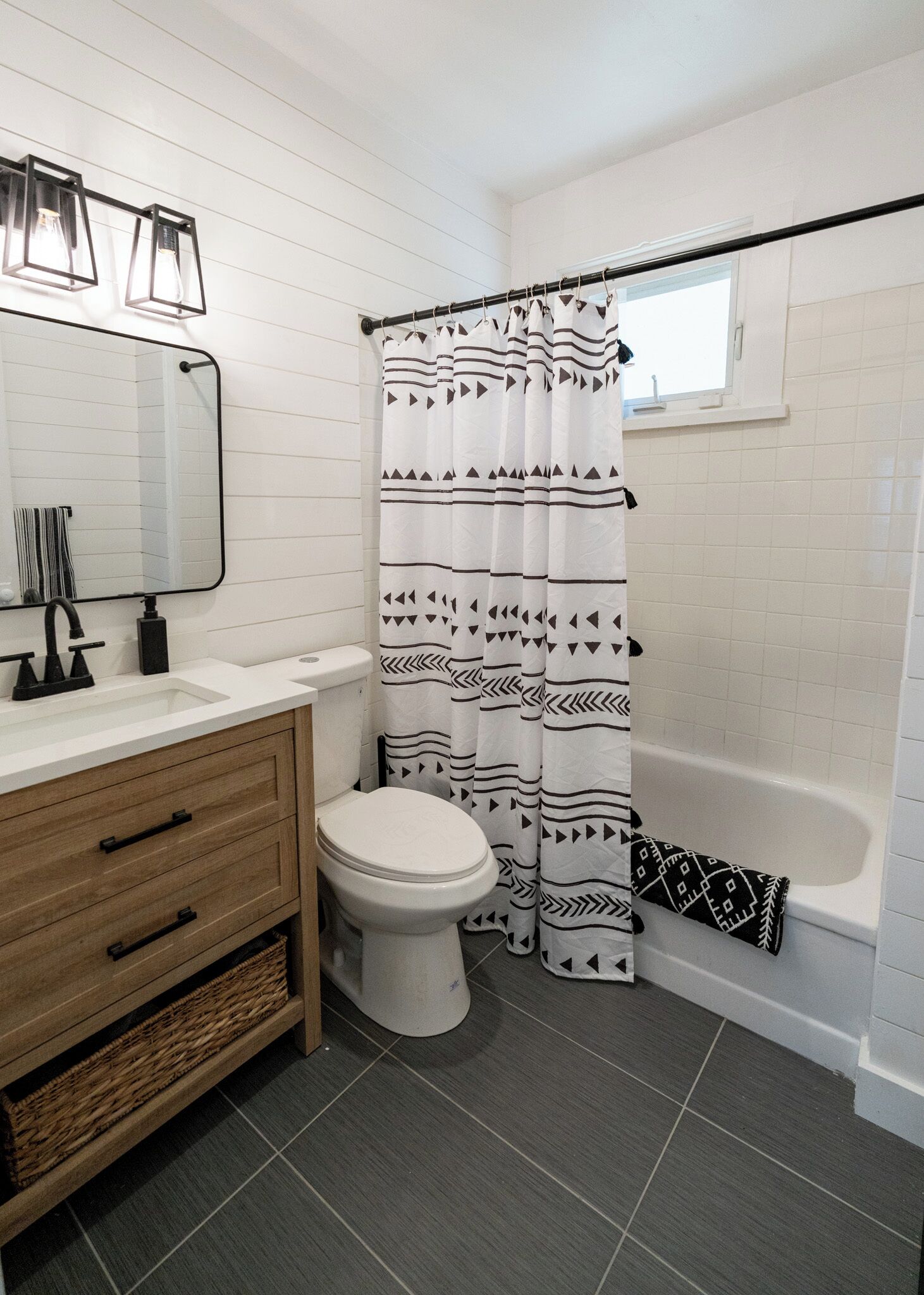 Une salle de bain avec des toilettes blanches, un meuble-lavabo en bois avec un miroir, un rideau de douche à motifs noir et blanc et une baignoire.