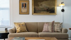 A living room with a beige sofa, patterned pillows, a framed painting, and a table with a book and keys. - Where We Roam Cottage - Walk to Downtown & Lake (Coeur d'Alene)