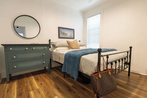 Un dormitorio con cama, cómoda, espejo, un cuadro y ventana con persianas.