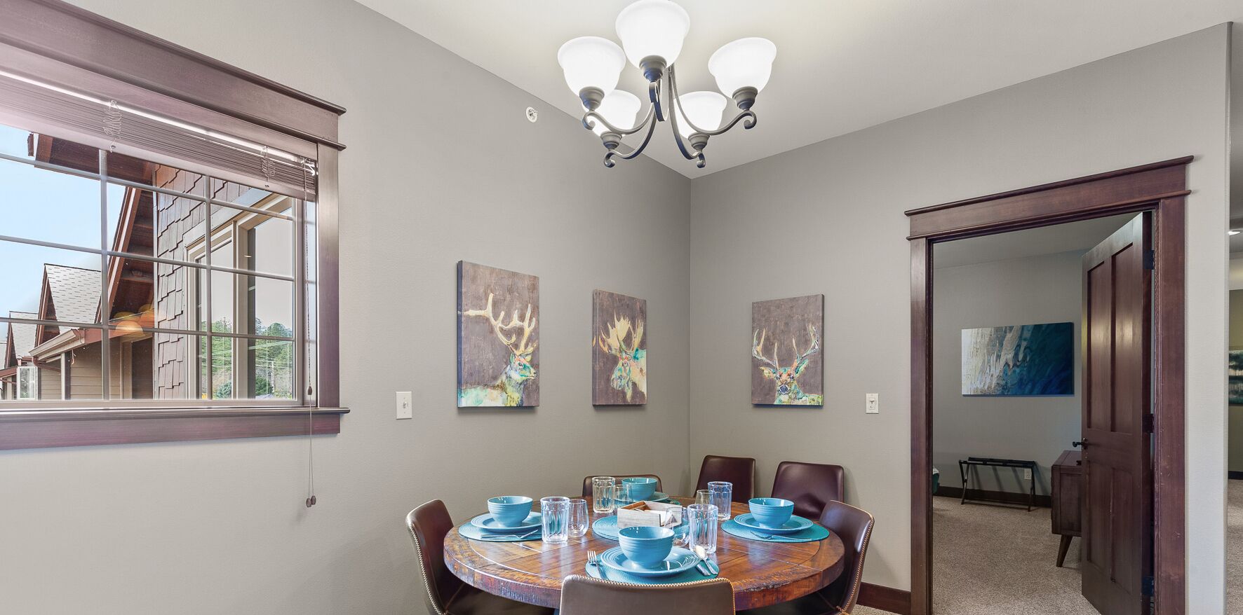 Een eetkamer met een ronde tafel voor zes personen, voorzien van blauwe borden en glazen. Er hangen drie schilderijen met hertenmotieven aan de muur, en een kroonluchter boven de tafel.