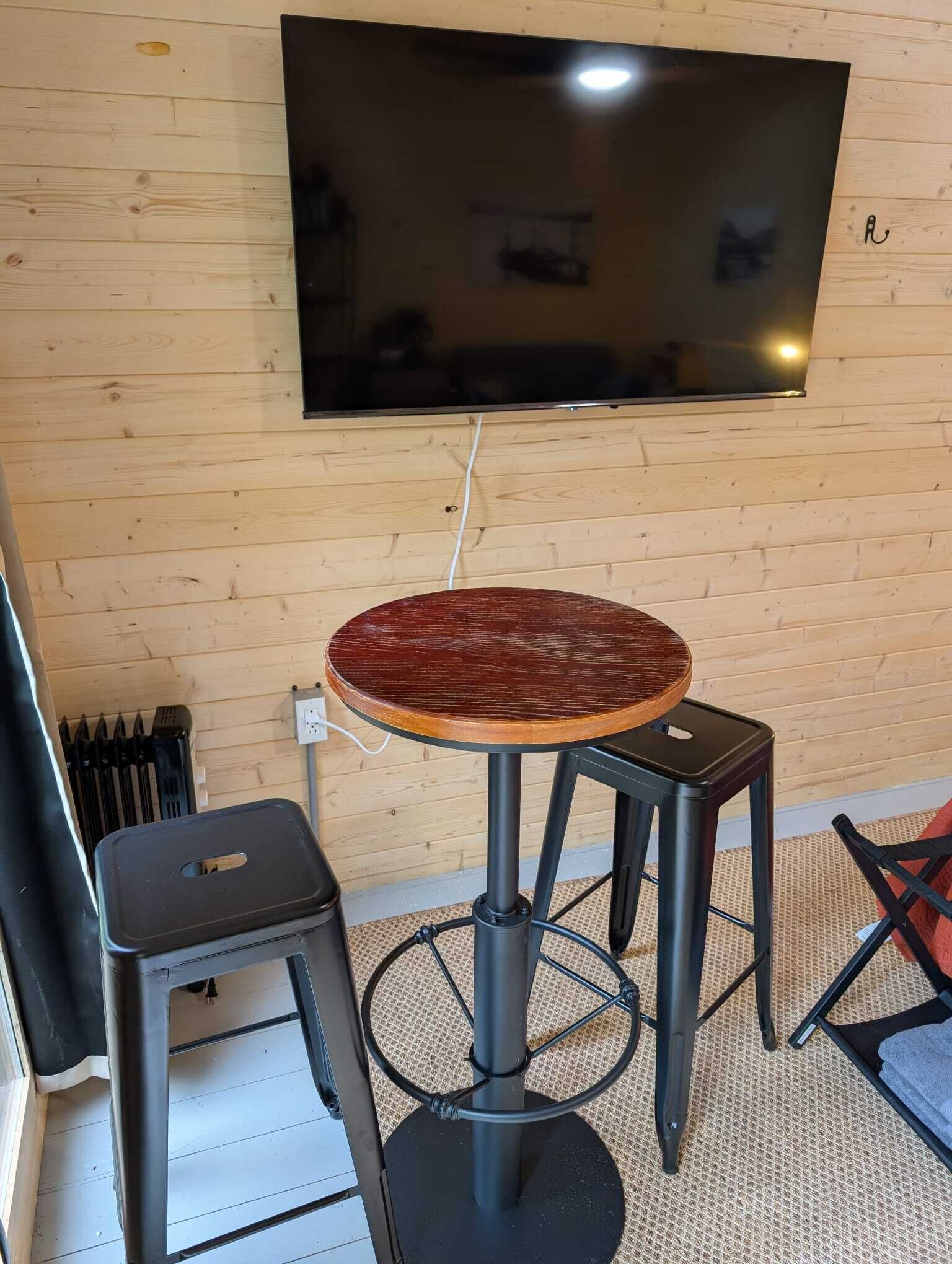 A room with a wooden wall, a round table with a dark wooden top, two black stools, and a mounted flat-screen TV.
