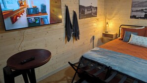 A cozy room with a bed, a TV, a small table, and a chair. There are pictures on the wall and a lamp on the nightstand.
