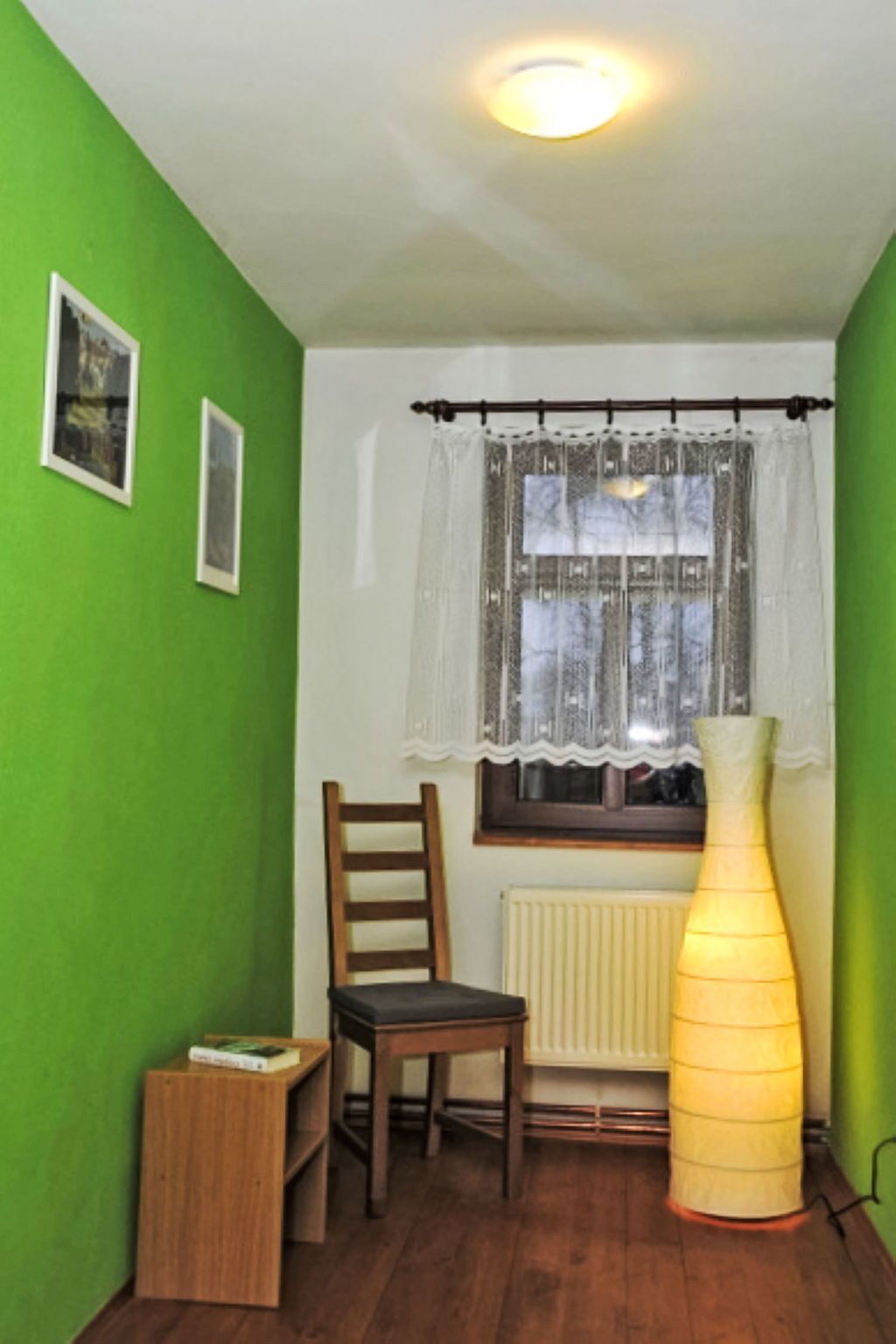 Et værelse med grønne vægge, en træstol, et lille træbord, en radiator og et vindue med gardin.