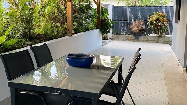 Eine Terrasse im Freien mit einem Tisch aus Glas und vier Stühlen, umgeben von Grünpflanzen und einem Fliesenboden.