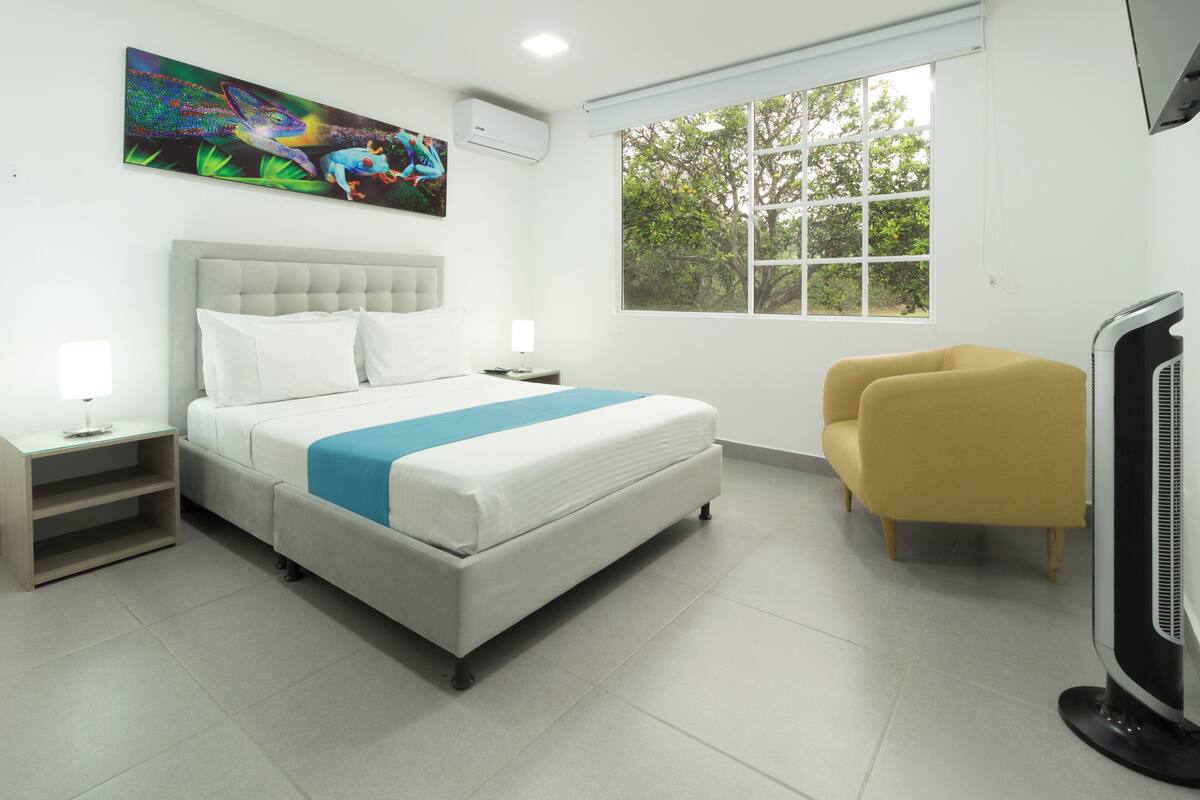 A modern hotel room with a bed, bedside tables, a flat-screen TV, a window with a view of trees, and a colorful artwork on the wall.