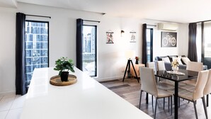 A modern hotel room with a large dining table, a seating area with chairs, a potted plant, and artwork on the walls.