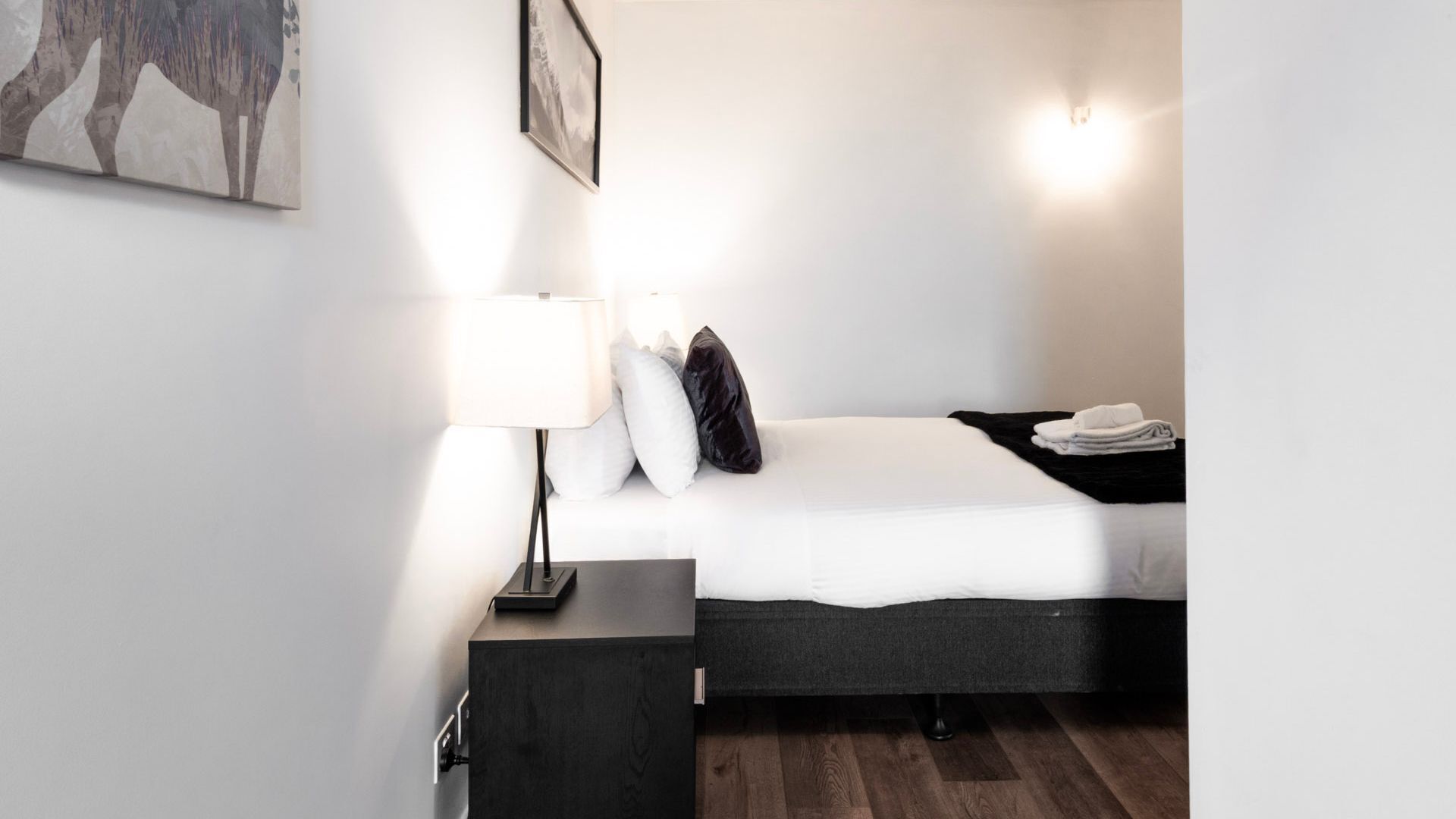 A modern hotel room with a bed, a nightstand, a lamp, and a wall-mounted light.