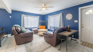 A living room with blue walls, a ceiling fan, a flat-screen TV, a brown leather sofa, a wooden coffee table, and a small table with a candle. - Dune's Edge Retreat Peaceful home near State park and trails (South Haven)