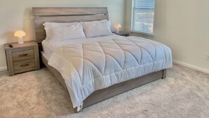 A bedroom with a bed, two bedside tables, a nightstand, a window with blinds, and a carpeted floor.