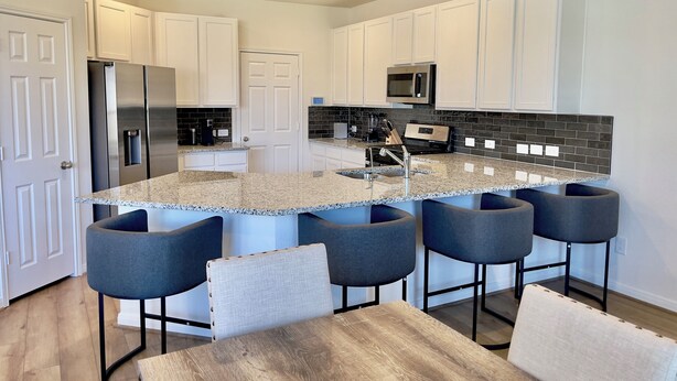 A modern kitchen with white cabinets, a granite countertop, a stainless steel refrigerator, a microwave, and a built-in oven. There are four bar stools and a wooden table.