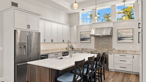 A modern kitchen with white cabinetry, stainless steel appliances, a large island with bar stools, and a view of palm trees through the windows. - Spectacular Direct access canal front beauty! (Sanibell)