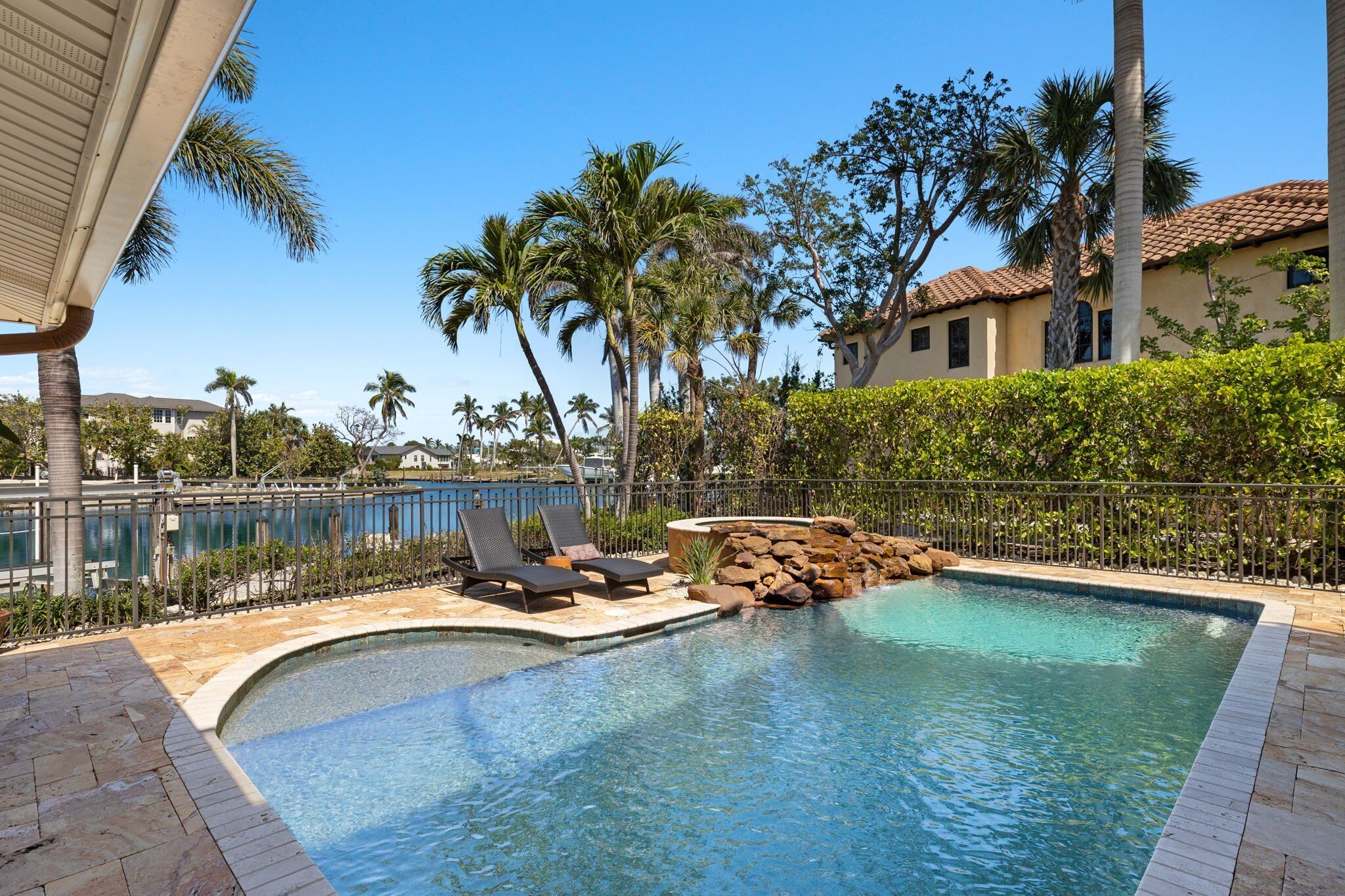A swimming pool with lounge chairs, surrounded by palm trees and a stone wall.