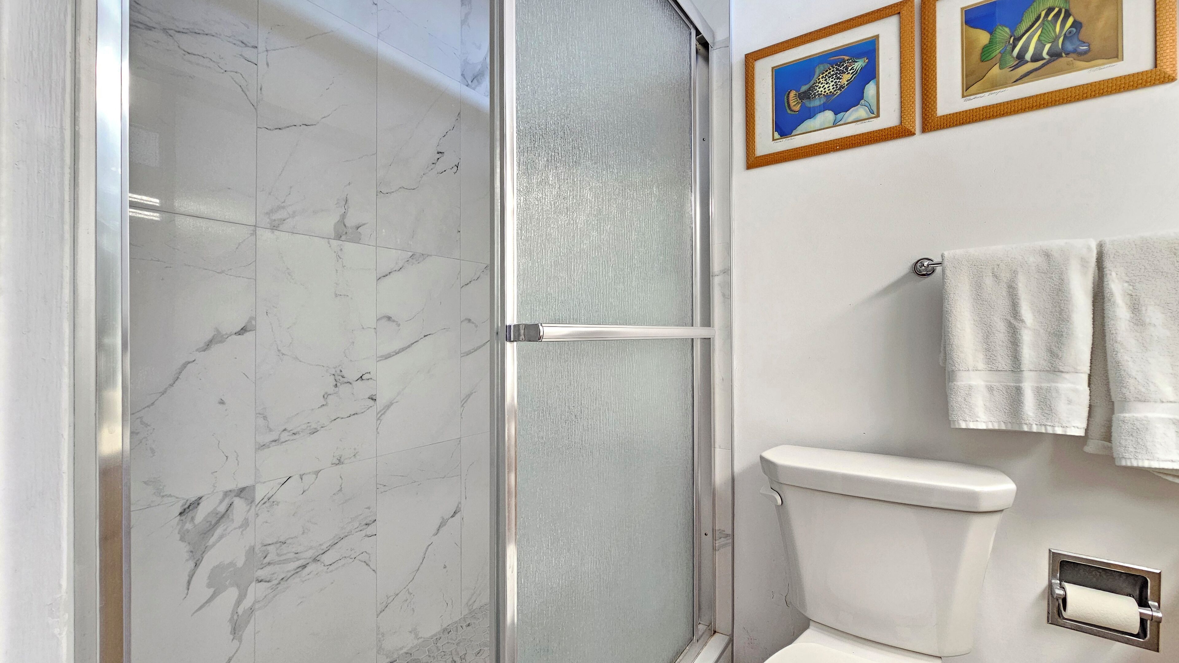 A bathroom with a marble shower enclosure, a toilet, and framed fish illustrations on the wall.