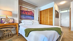 A neatly arranged bedroom with a bed, a bedside table with a lamp, a framed picture, and a painting on the wall. - Beachfront 8th Floor Sans Souci Studio - 30 Day Rental (Honolulu)