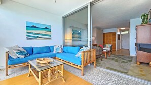 A living room with a blue sofa, a glass coffee table, and a wooden chair. There is a painting of a beach scene on the wall. - Beachfront 8th Floor Sans Souci Studio - 30 Day Rental (Honolulu)