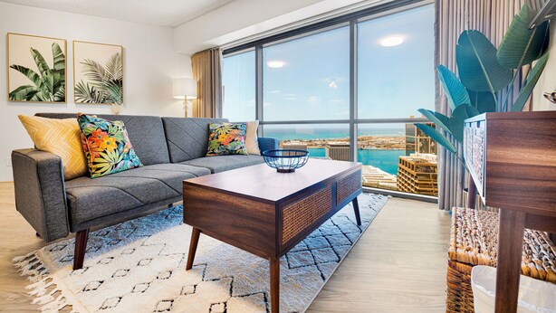 A modern living room with a gray sofa, colorful cushions, a wooden coffee table, and large windows offering a view of the ocean.