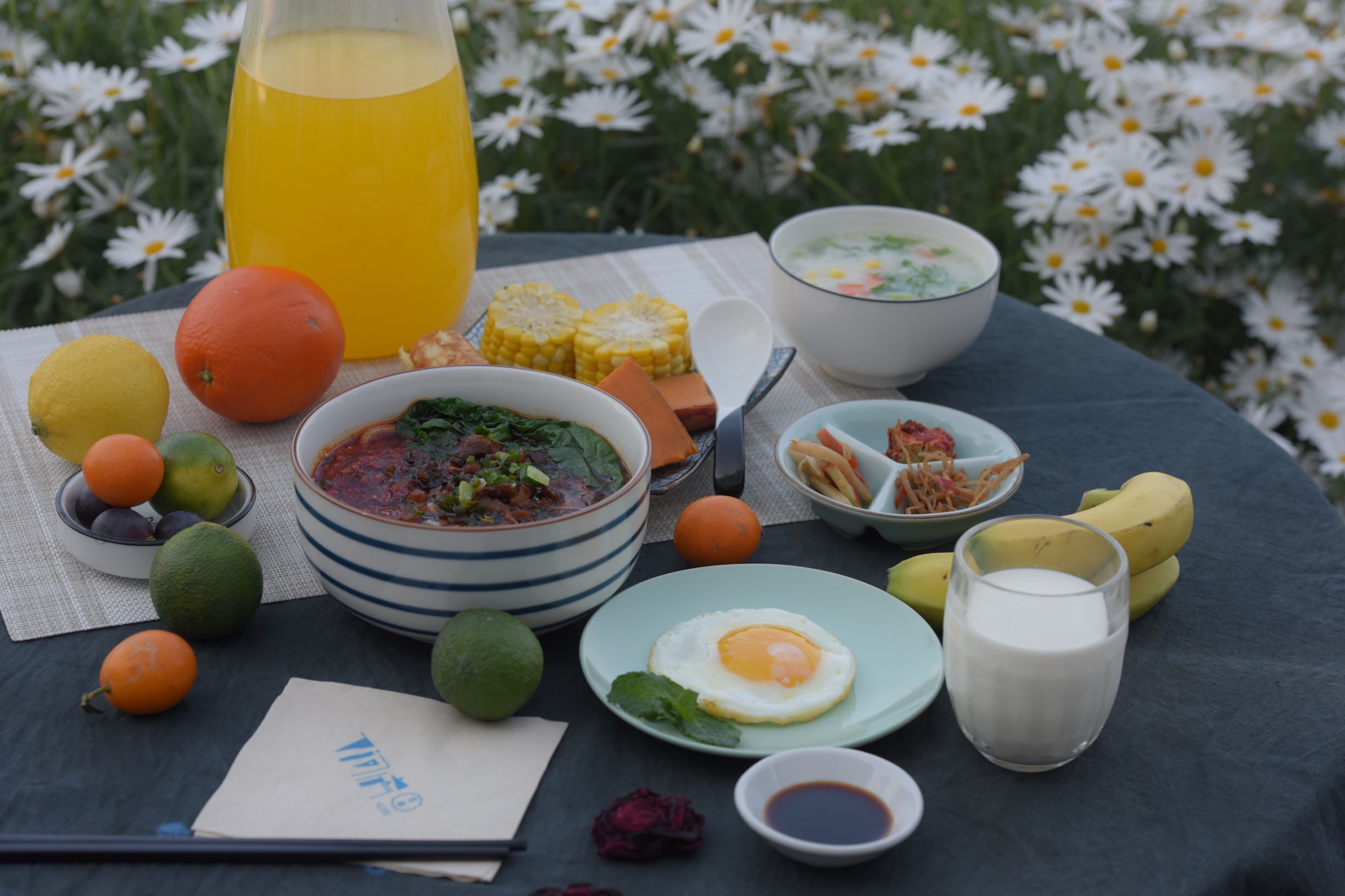 Una mesa servida con diversos platos, incluyendo sopa, huevos y frutas, con una botella de jugo de naranja y un tazón de sopa al fondo.