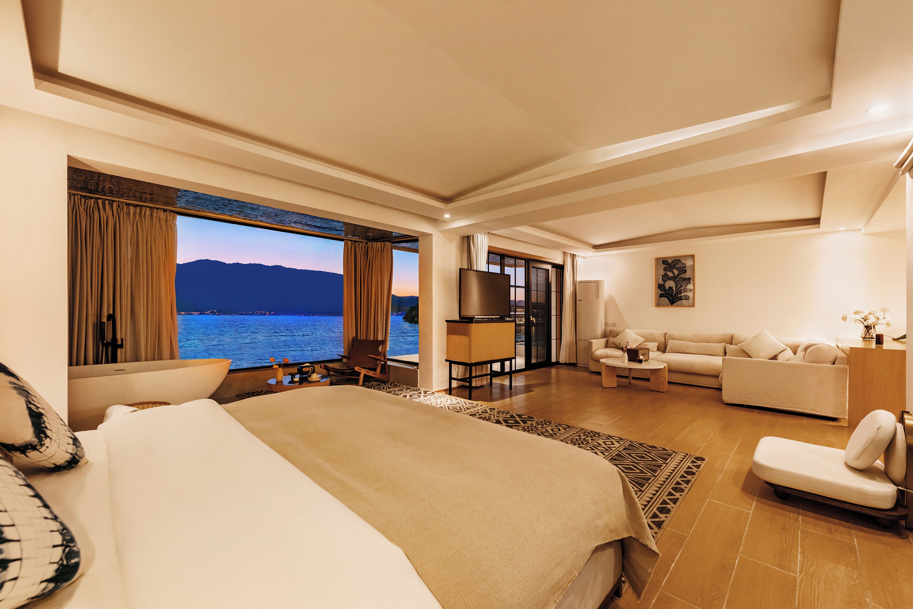 A spacious hotel room with a large bed, a sofa, a chair, and a TV. There is a view of the sea through the window.