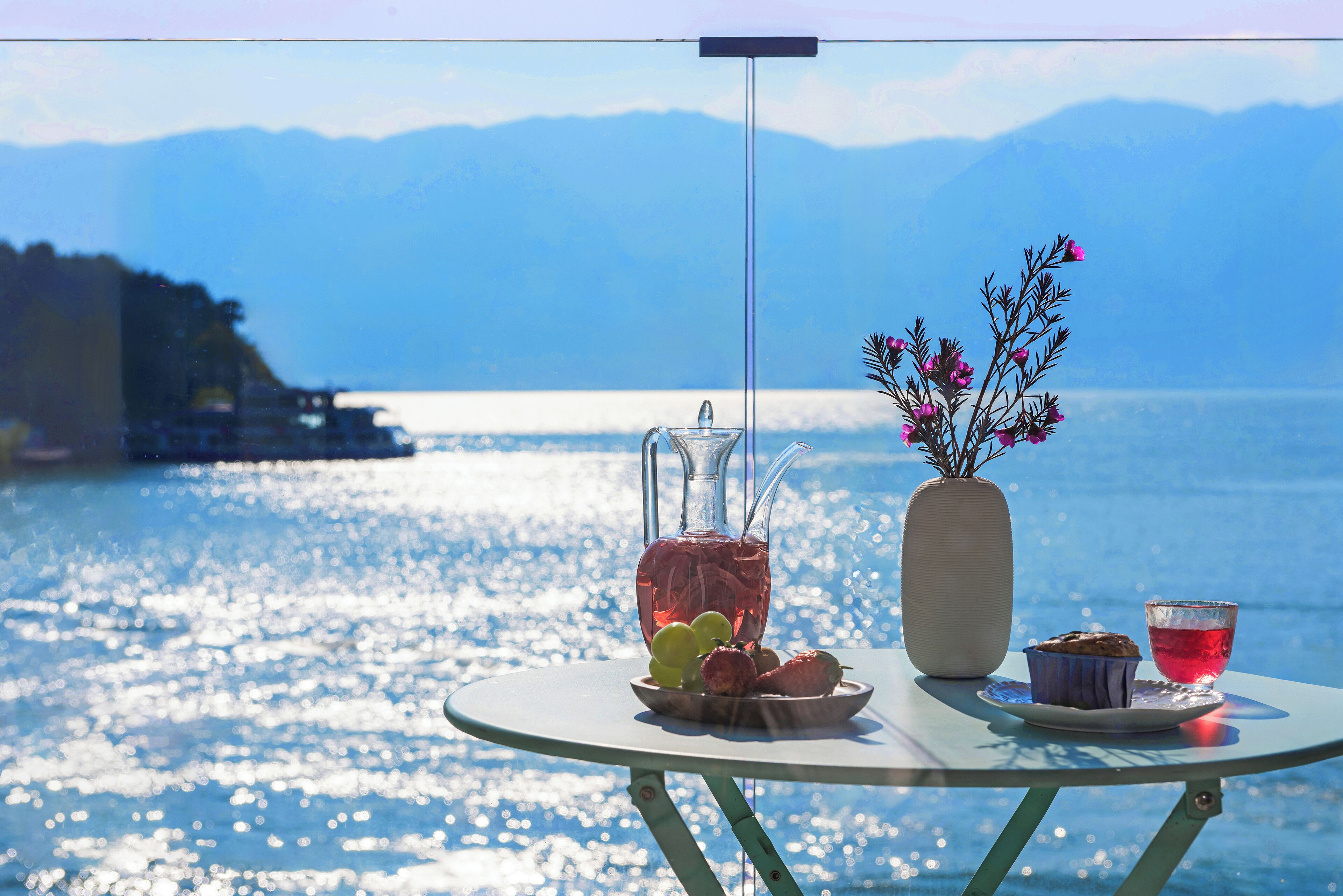 A table with a pitcher, a vase with flowers, a plate with fruits, and a cup of tea, set against a scenic lake view.