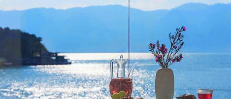 A table with a pitcher, a vase with flowers, a plate with fruits, and a cup of tea, set against a scenic lake view.