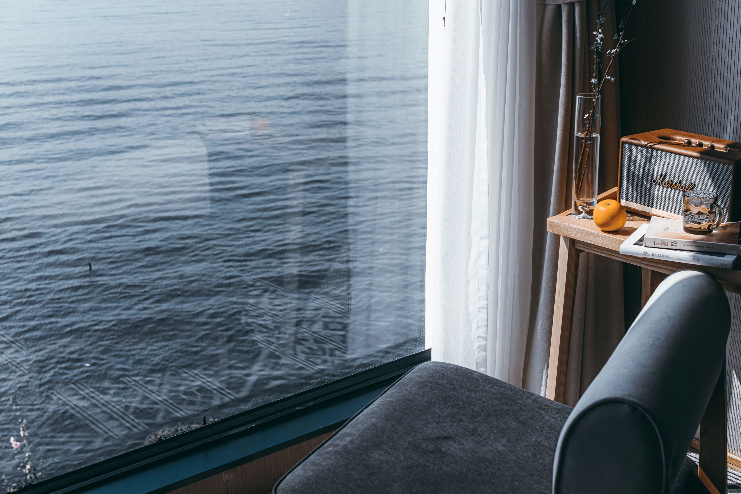 Une pièce avec vue sur la mer, visible par une fenêtre, une petite table avec une radio ancienne, un coussin et un vase en verre rempli de fleurs.