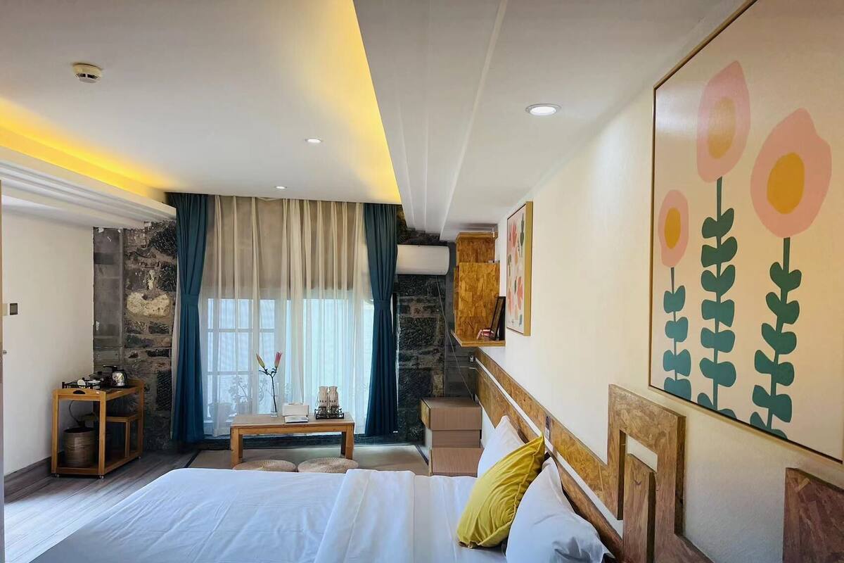 Un dormitorio con una cama grande, cabecera de madera, mesita de noche, una pequeña mesa y ventana con cortinas.