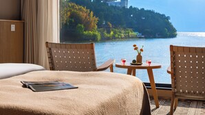 Quarto de hotel com cama, duas cadeiras, uma mesinha com um vaso e bebidas, e vista para um lago e montanhas.