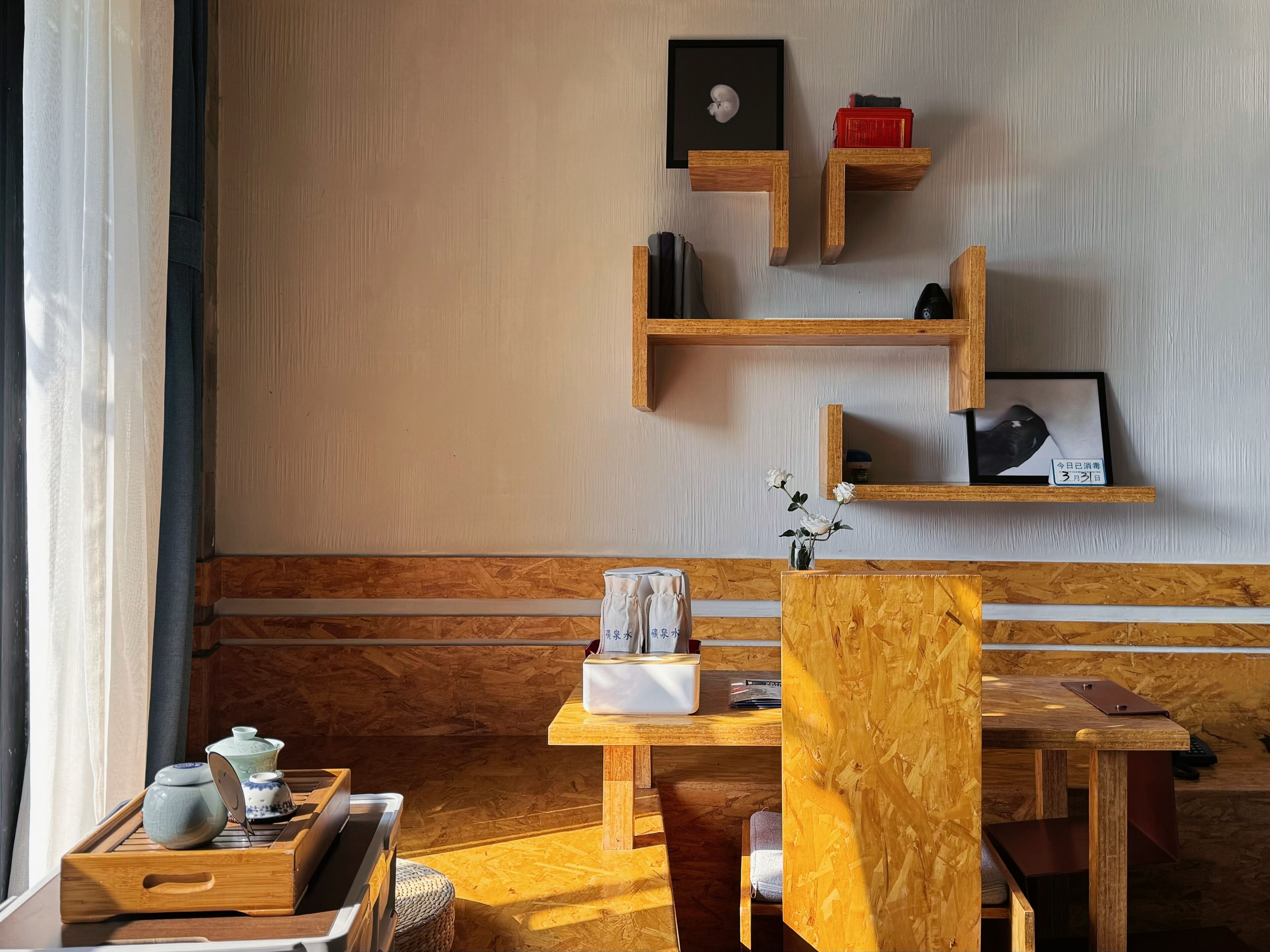 Una habitación con muebles de madera, un banco de madera, una pequeña mesa con un florero y una estantería con un cuadro enmarcado y una caja roja.