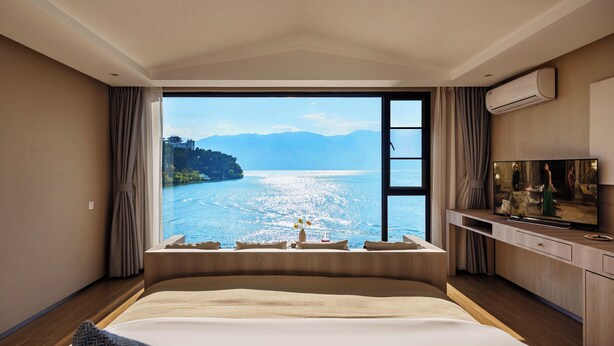 Quarto de hotel com uma cama grande, televisão e vista para um lago e montanhas.
