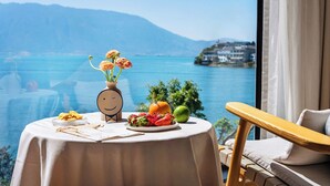 Uma mesa com uma travessa de frutas e um vaso de flores, uma cadeira de madeira e vista para um lago e montanhas.