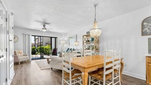 Amplia sala de estar con una mesa de comedor de madera, sillas blancas, un ventilador de techo y un ventanal con vistas al exterior.