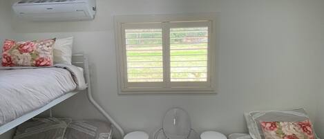 A bedroom with two bunk beds, a ceiling fan, an air conditioning unit, a window with a view of greenery, and a small bathroom with a toilet and sink.