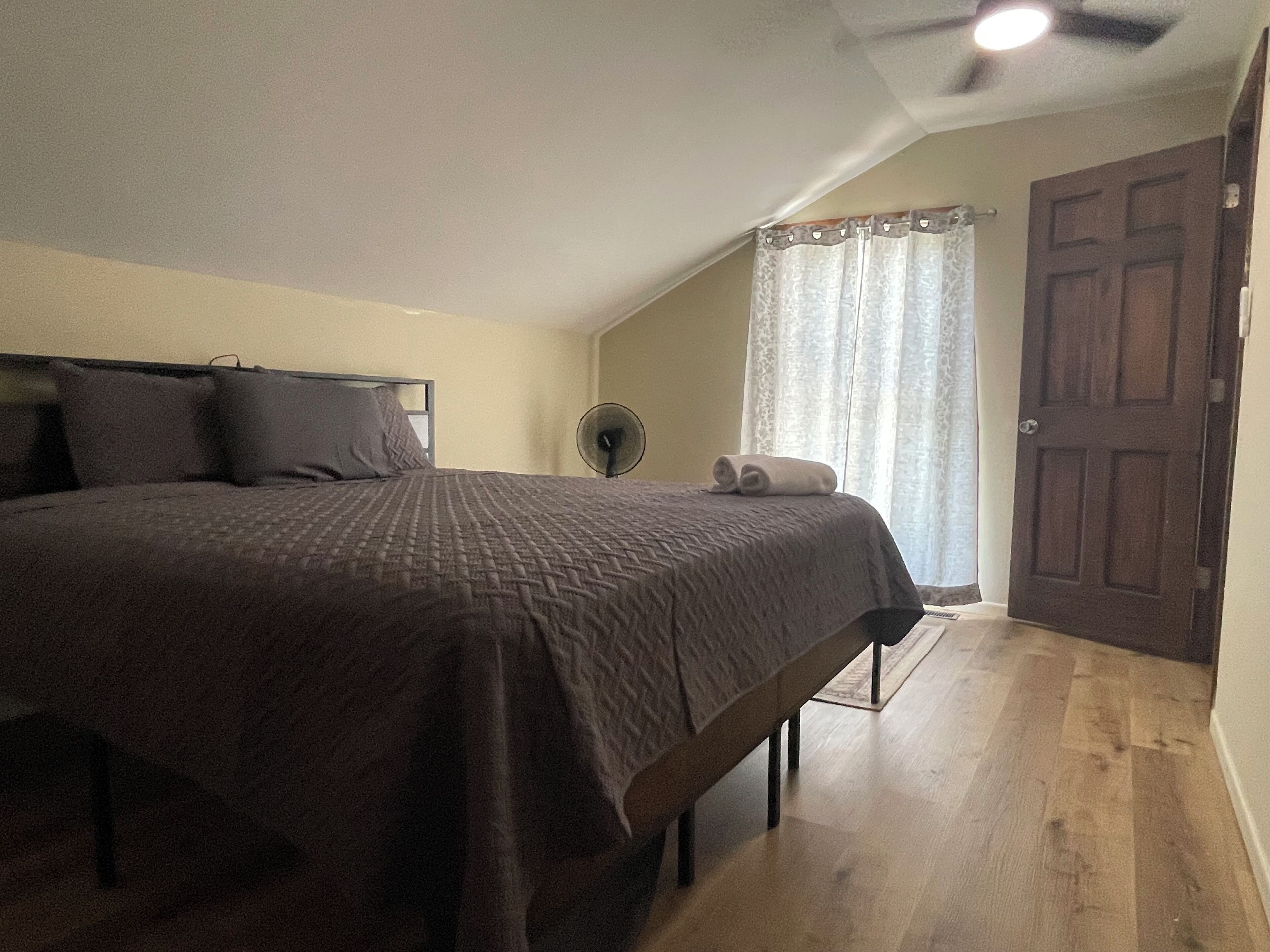 A bedroom with a large bed, a ceiling fan, a window with curtains, and a wooden door.