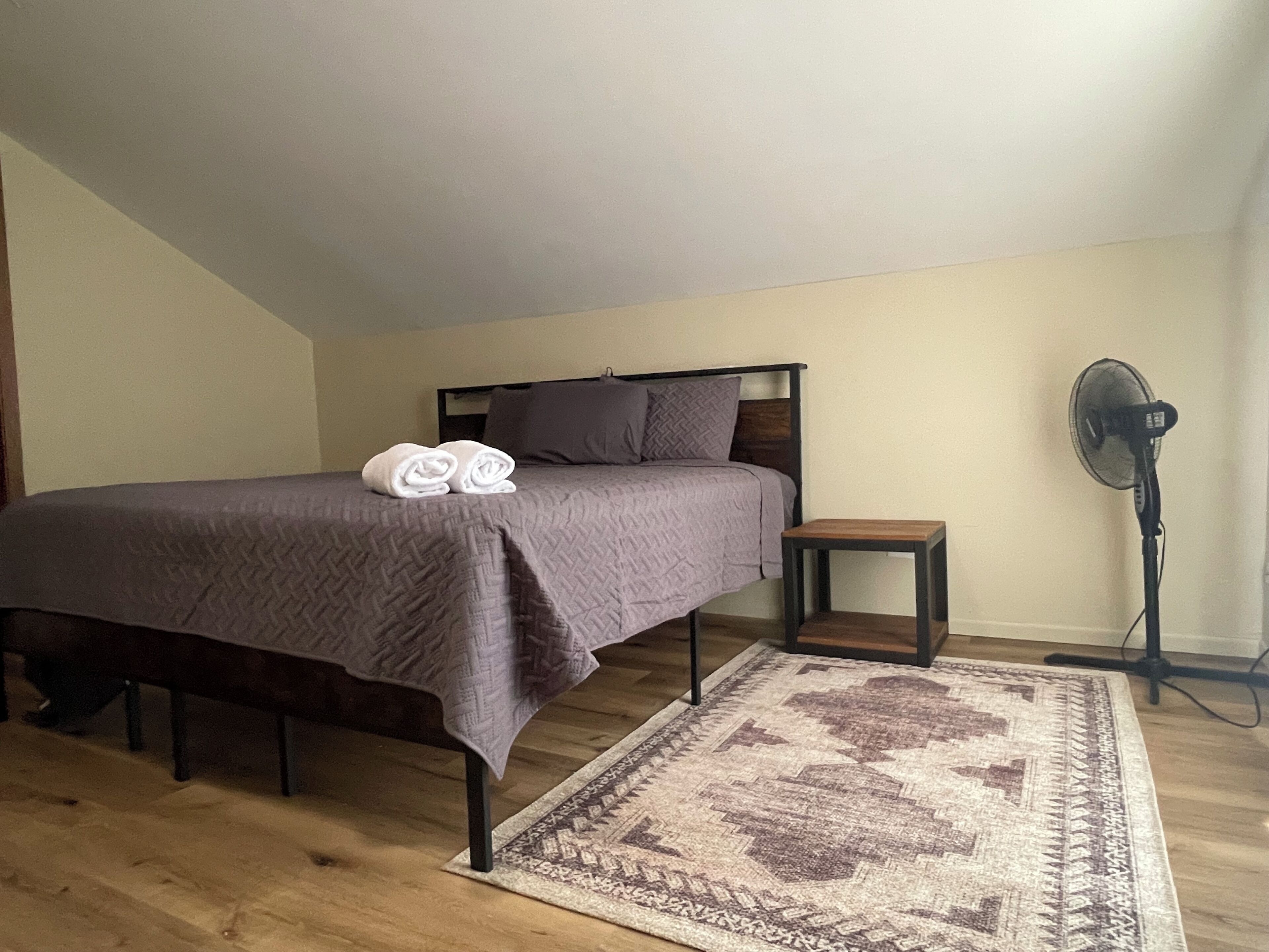 Een slaapkamer met een bed, nachtkastje, ventilator en een kamerkleed met patroon.