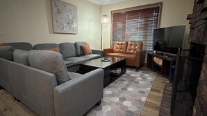 Een woonkamer met een grijze bank, een bruine leren fauteuil, een salontafel en een televisie.