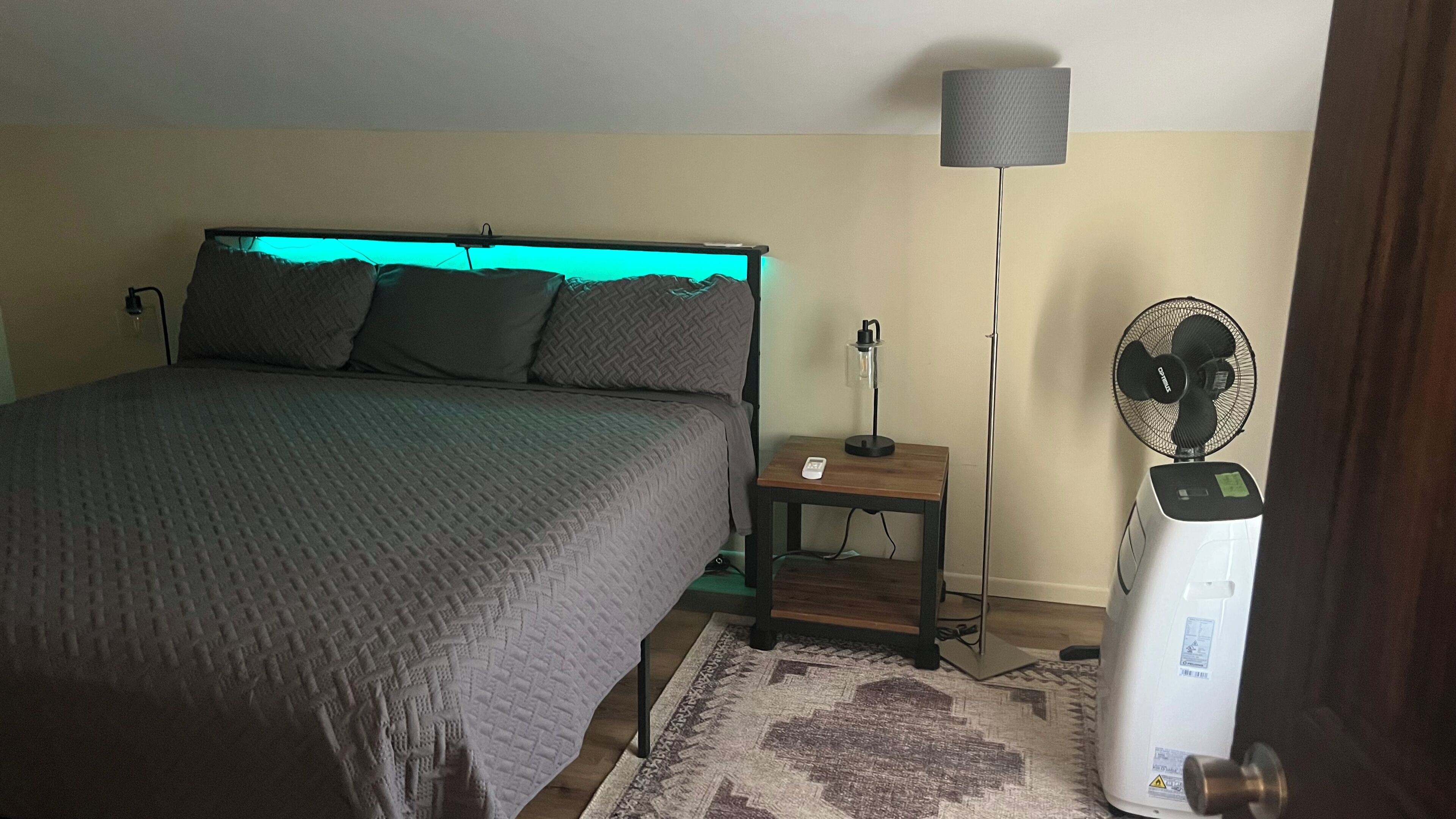A bedroom with a bed, bedside table, lamp, and a standing fan.