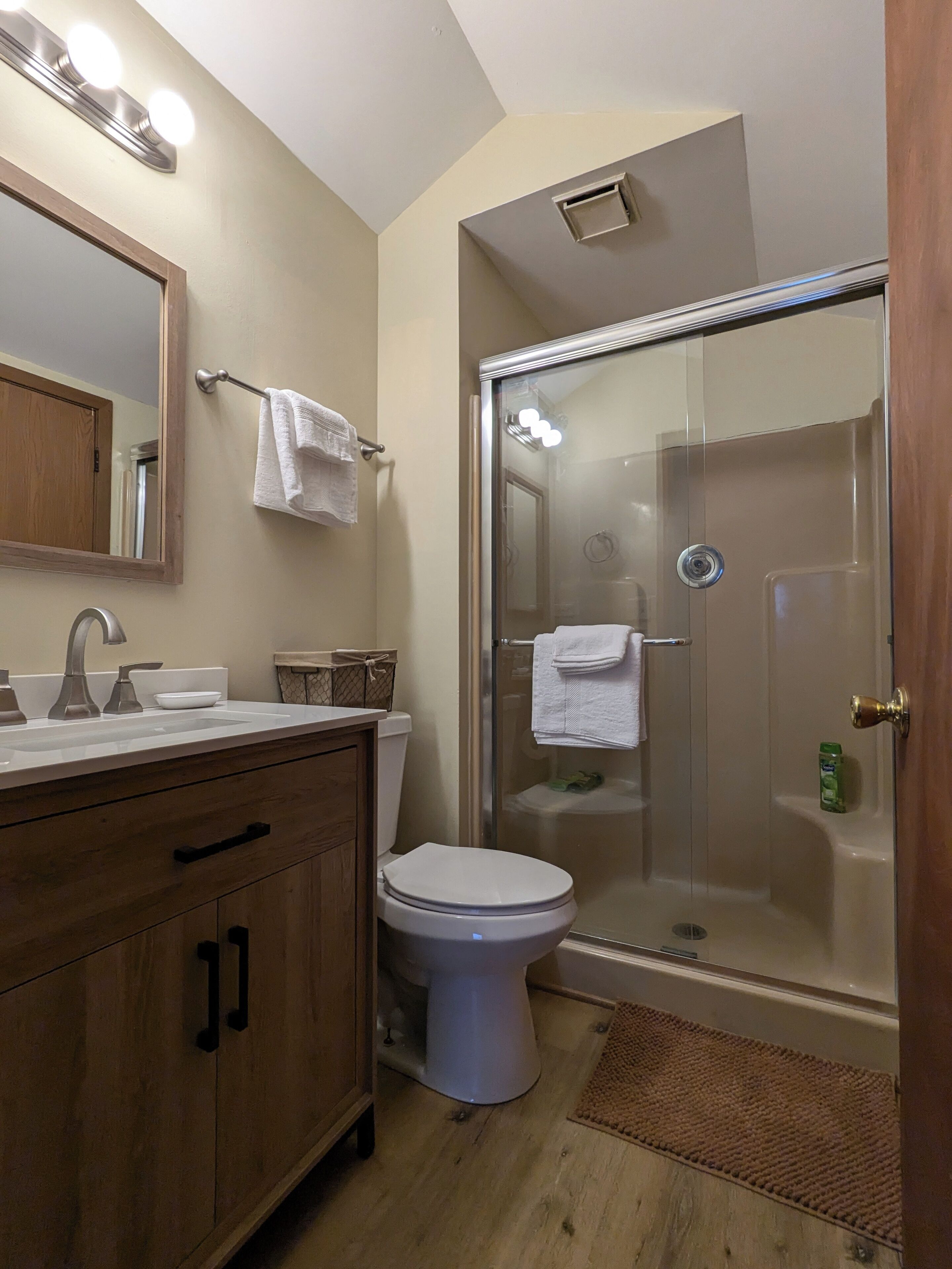 Un baño con tocador de madera, inodoro blanco, ducha con mampara de vidrio y espejo con iluminación superior.