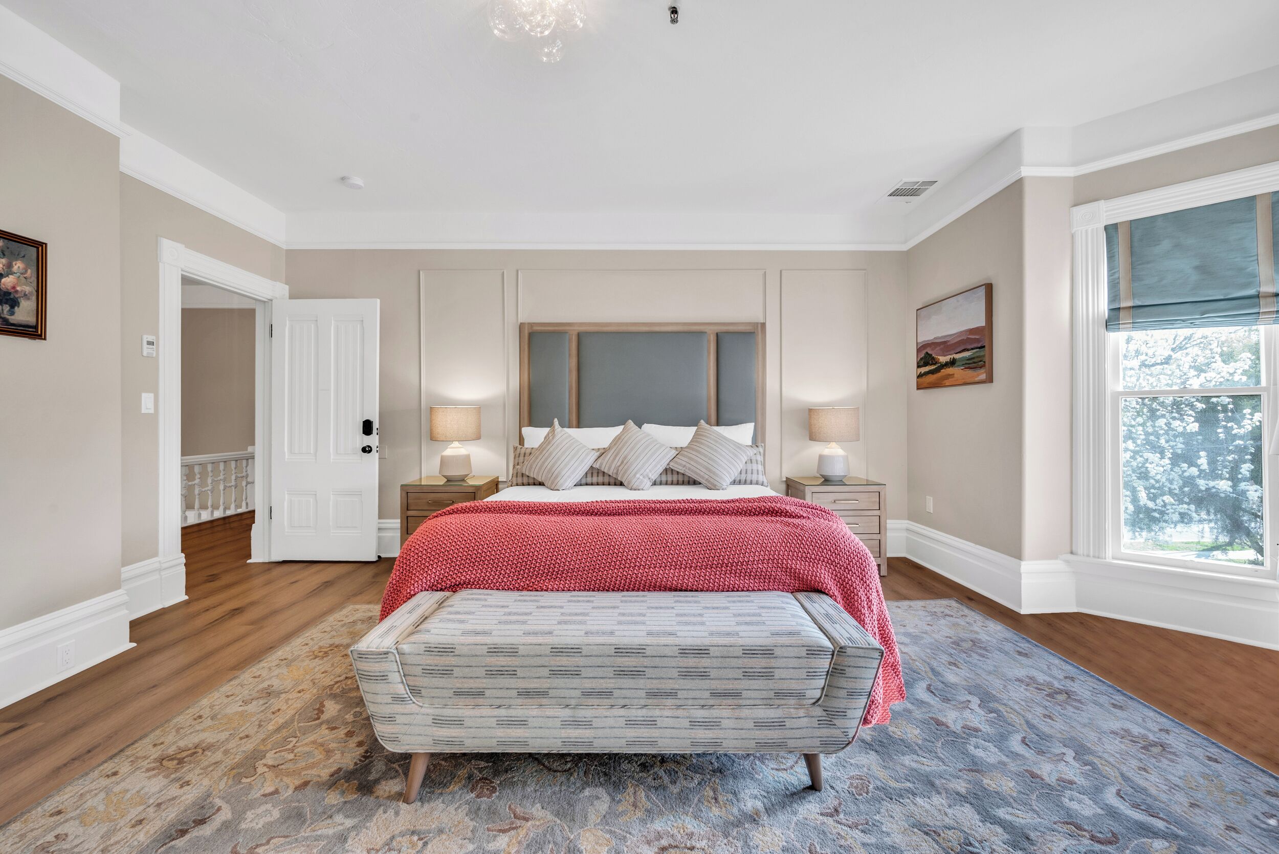 Een slaapkamer met een bed, nachtkastjes, een bankje, een groot raam en een deur die naar een andere kamer leidt.