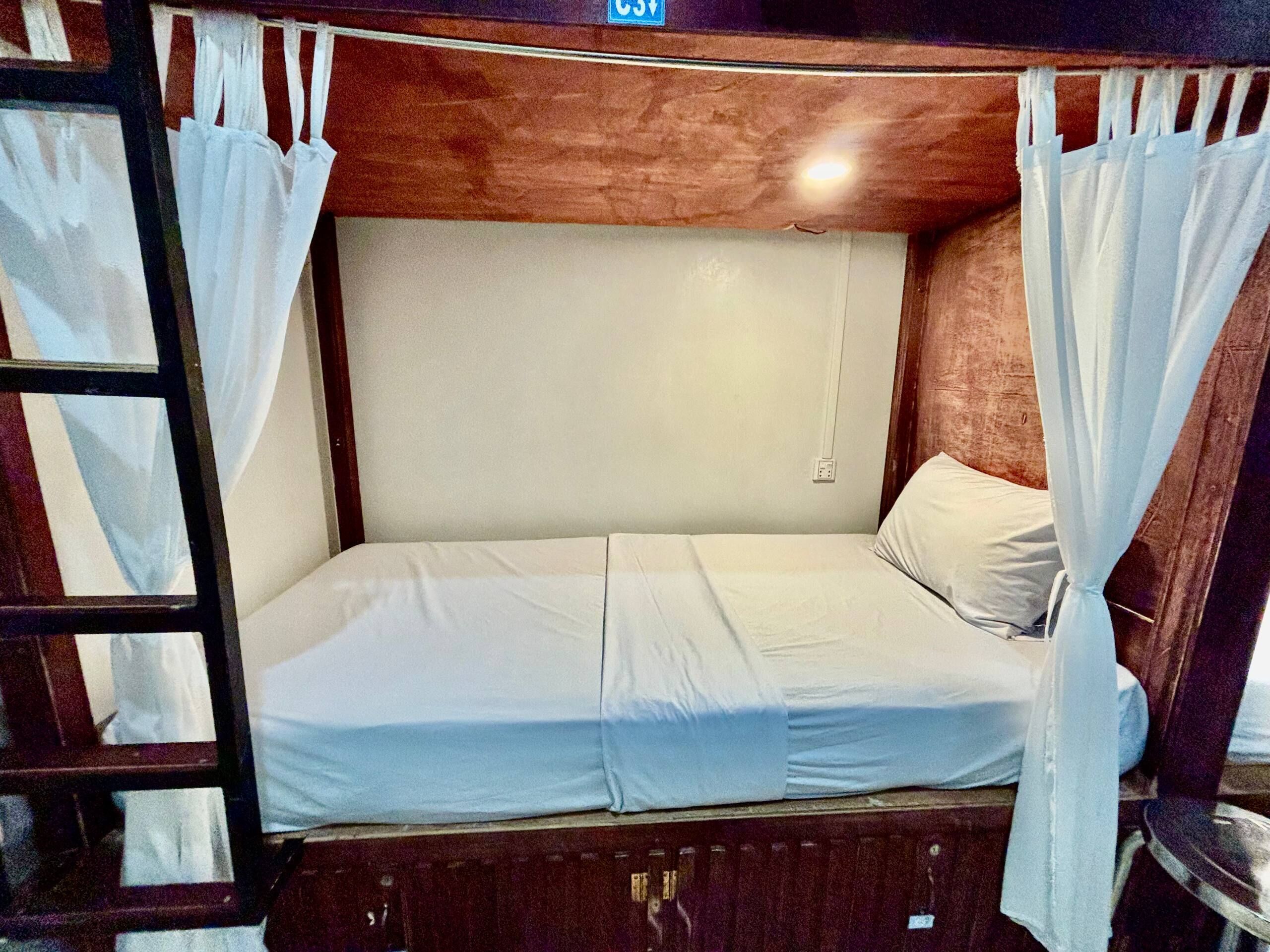Una cama individual en literas con ropa de cama blanca, cabecera de madera y cortinas blancas.