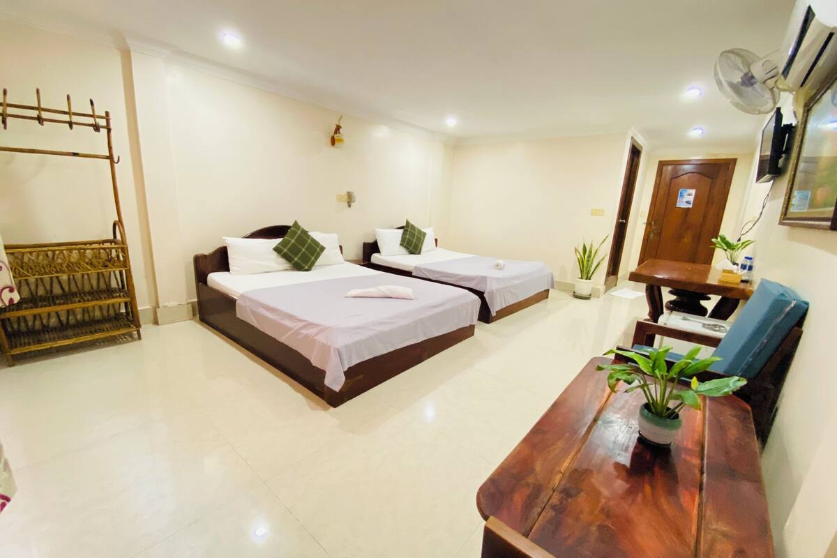 Una habitación de hotel con dos camas, un escritorio de madera, una silla, un ventilador y una pequeña planta en maceta.