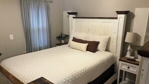 A bed with a quilted white bedspread, a wooden headboard, and a bedside table with a lamp and remote control. - Cottage on Main st- Low Cleaning Fee (Saint Charles)